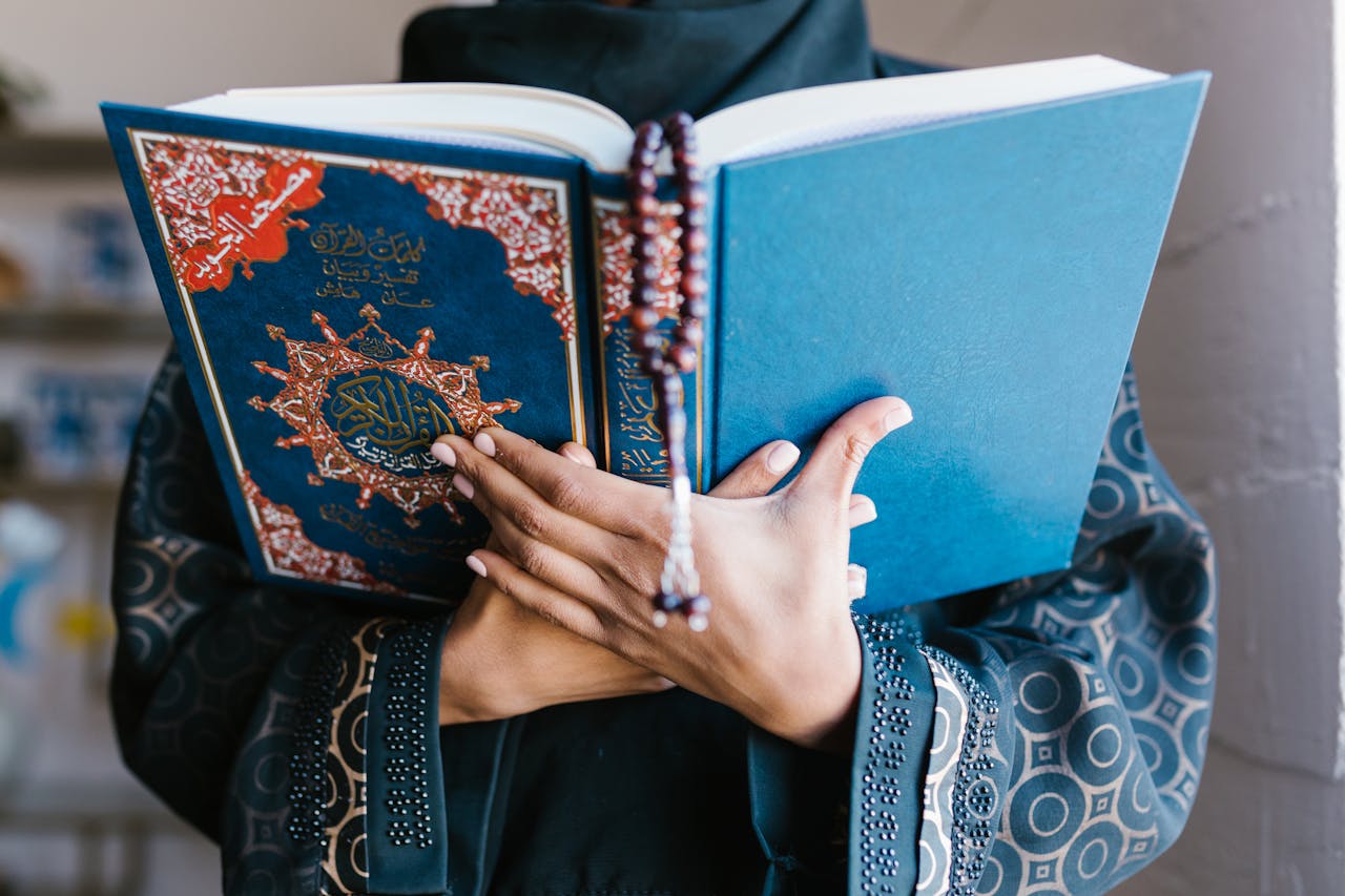 A Muslim woman in traditional attire reading the Quran indoors, holding prayer beads in a peaceful setting.